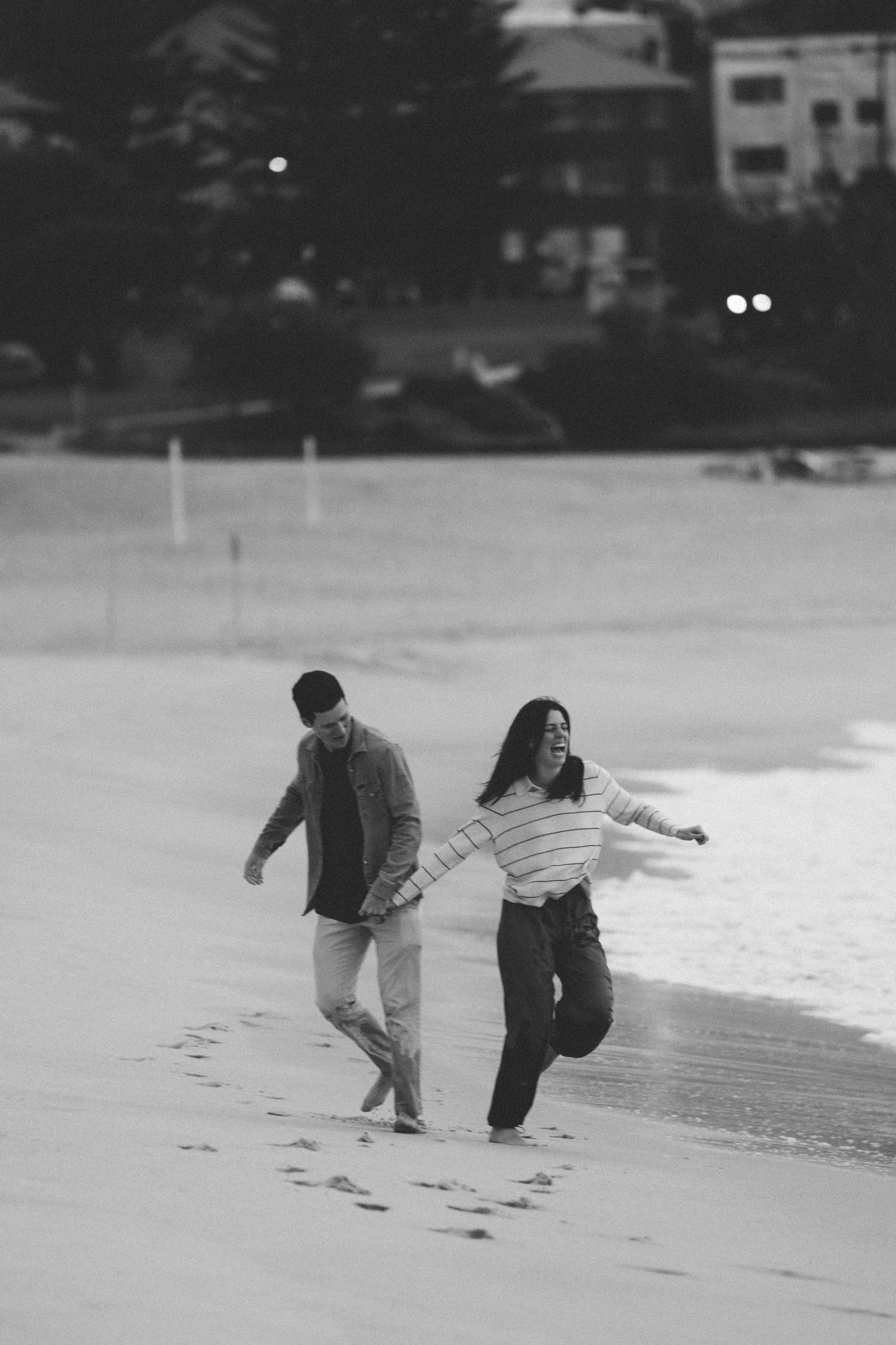 A couple running barefoot along the shoreline, laughing, in black and white.