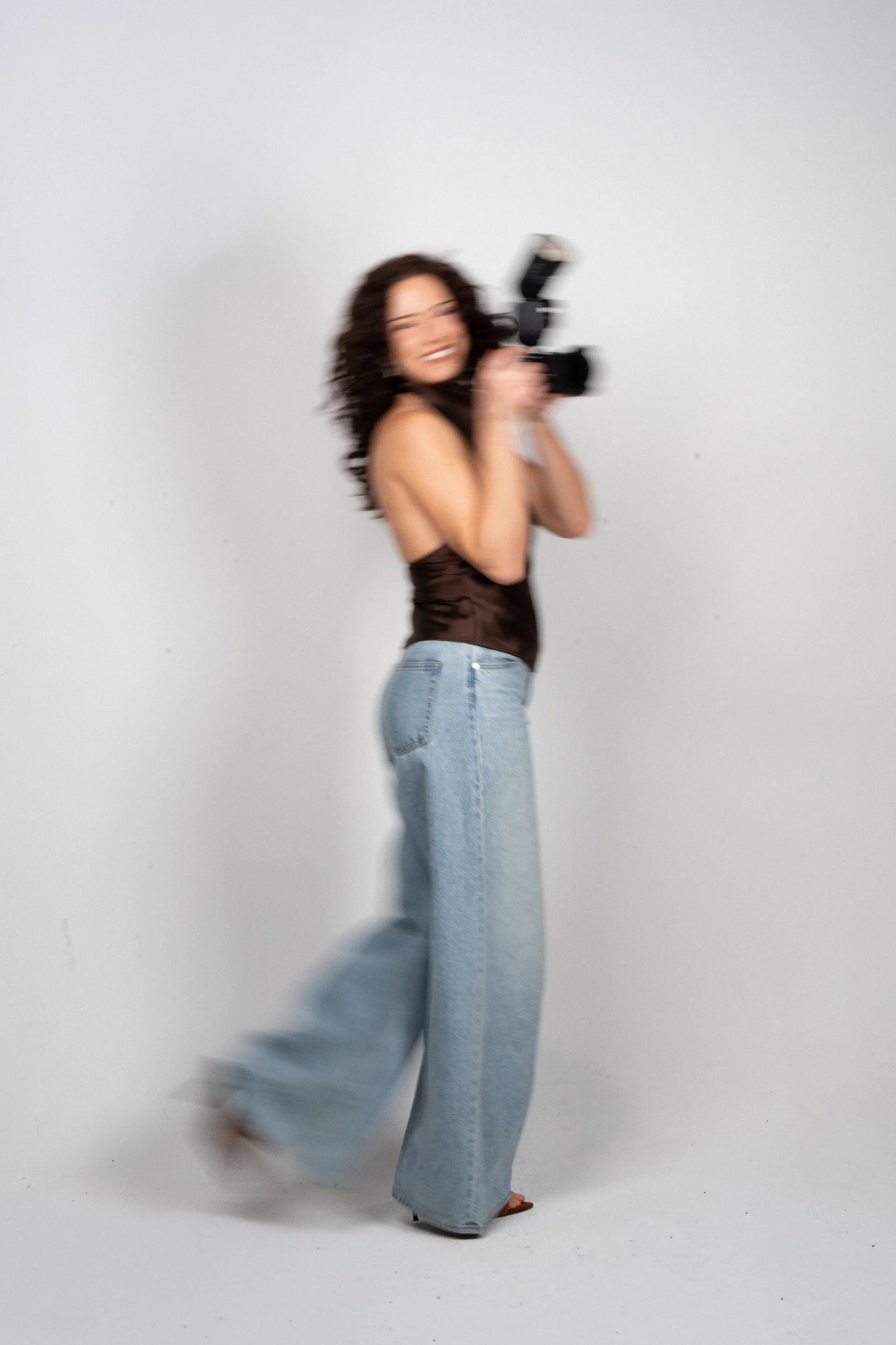 Flora mid-motion in a brown satin top and wide-leg jeans, camera raised and blurred, against a white studio backdrop.
