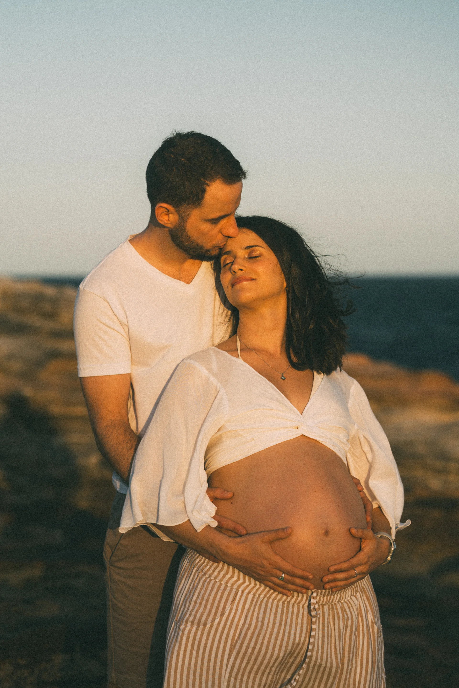 A couple at golden hour, his lips to her forehead, hands cradling the bump