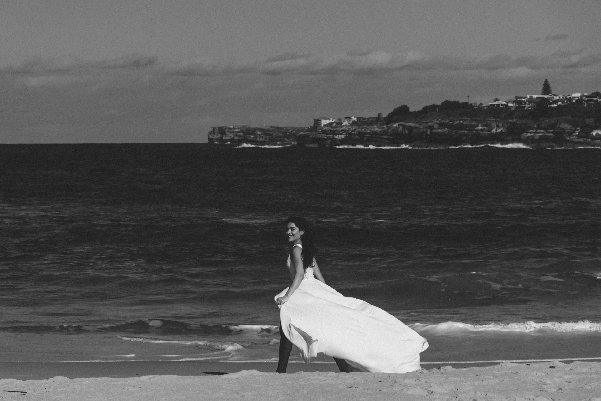 A bride standing on the sand by the water, in black and white, Sydney